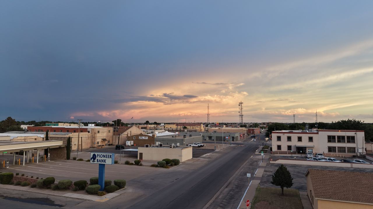 Aerial drone photo of downtown Carlsbad, New Mexico at sunset: the main street and Pioneer Bank sign below a deep blue sky glowing amber and coral on the horizon