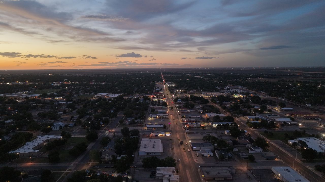 Drone photo looking down Canal Street in Carlsbad, NM at sunset, with lit businesses and car headlights leading to an orange horizon