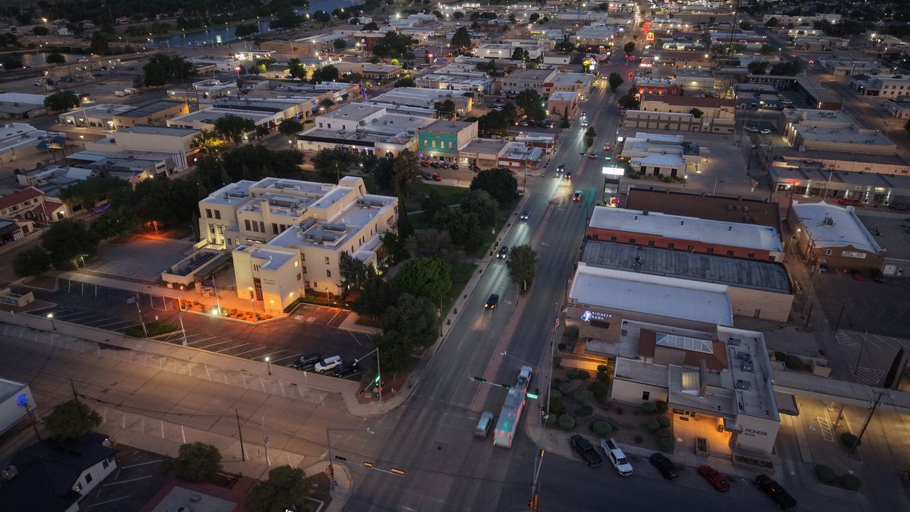 Twilight aerial of downtown Carlsbad, NM, showing a lit government building, Canal Street traffic, and the river in the distance