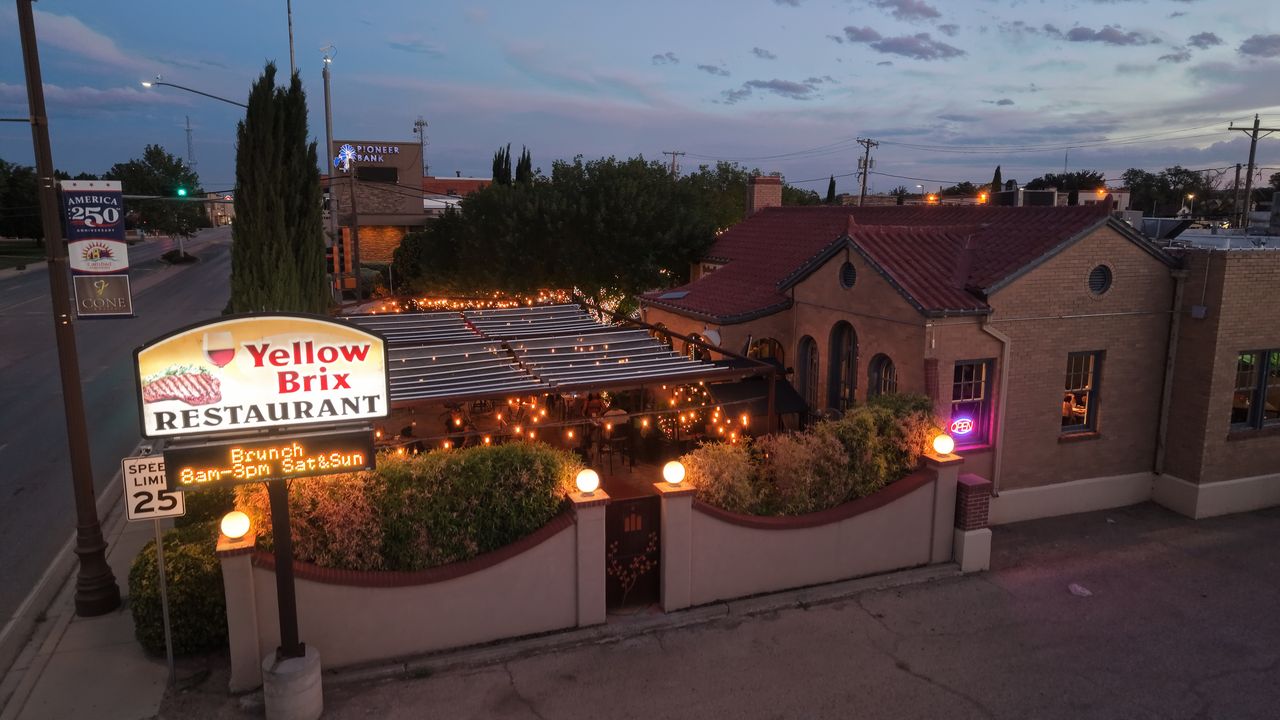 Low-altitude drone photo of Yellow Brix Restaurant on Canal Street in Carlsbad, NM at twilight, with its lit sign and string-lit patio